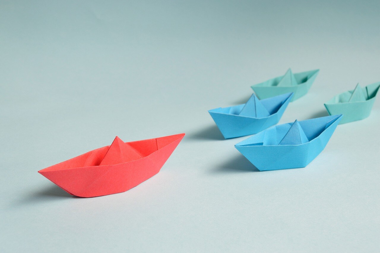 A red paper boat pulls ahead of blue and green paper boats. 